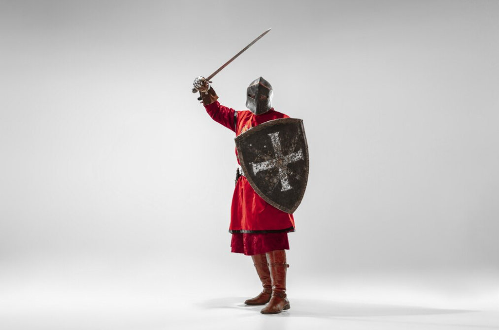 A knight in red tunic holding a sword and shield poses against a white wall